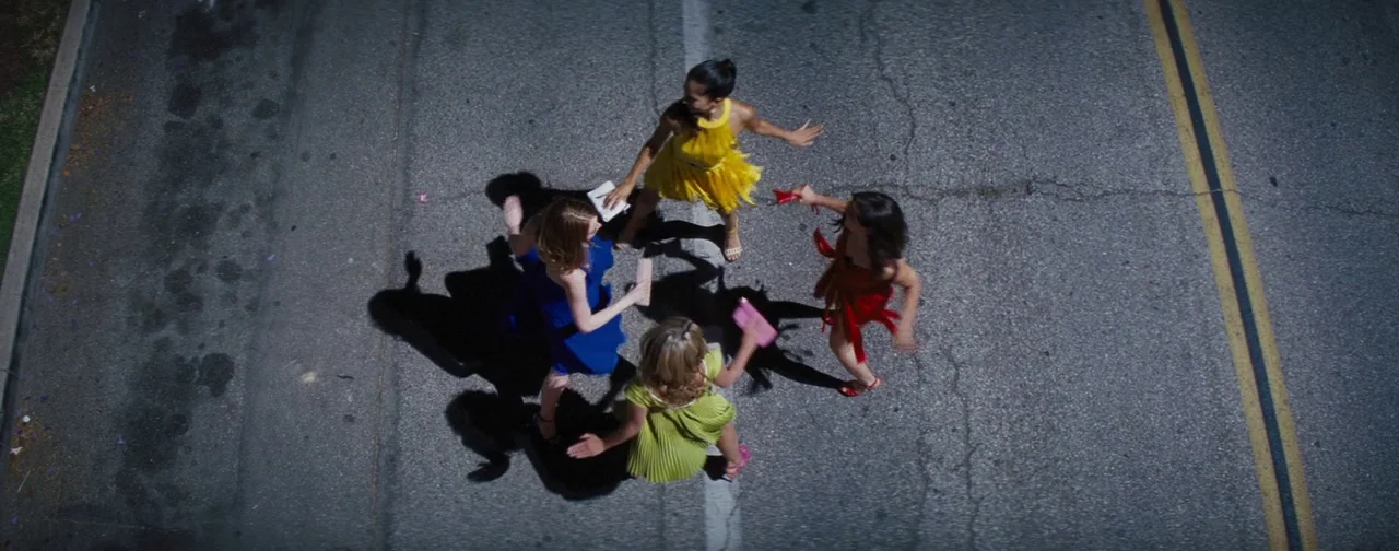 La La Land (2016) movie still: wide — Four women stand in a circle on a grey asphalt road under direct sun. Each woman wears a dress of a distinct…