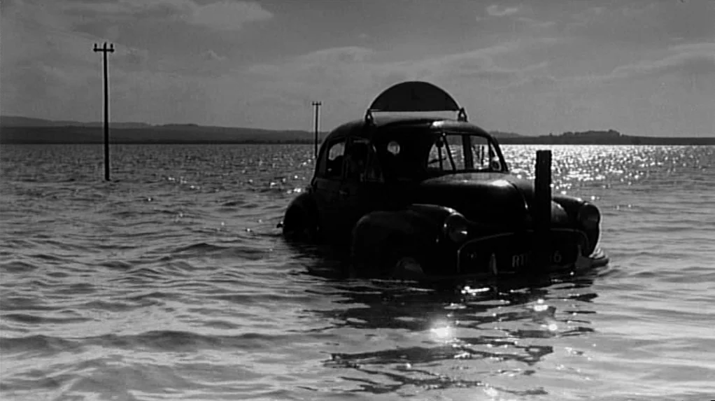 Cul-de-sac (1966) movie still: wide — A dark vehicle rests in the middle of a vast, flooded expanse under a cloudy sky. The car is partially…