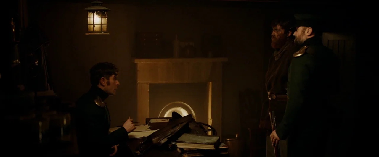 Black ’47 (2018) movie still: wide — A dark, dimly lit cabin features a stone hearth and a heavy wooden table. One man sits writing at the desk…