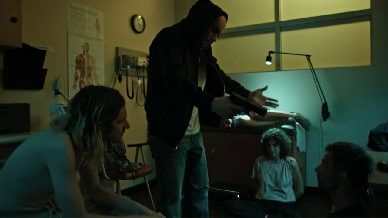 Bullet Proof (2022) movie still: medium — A clinical medical room features an anatomical chart on the wall and exam tables. A man holding a handgun…
