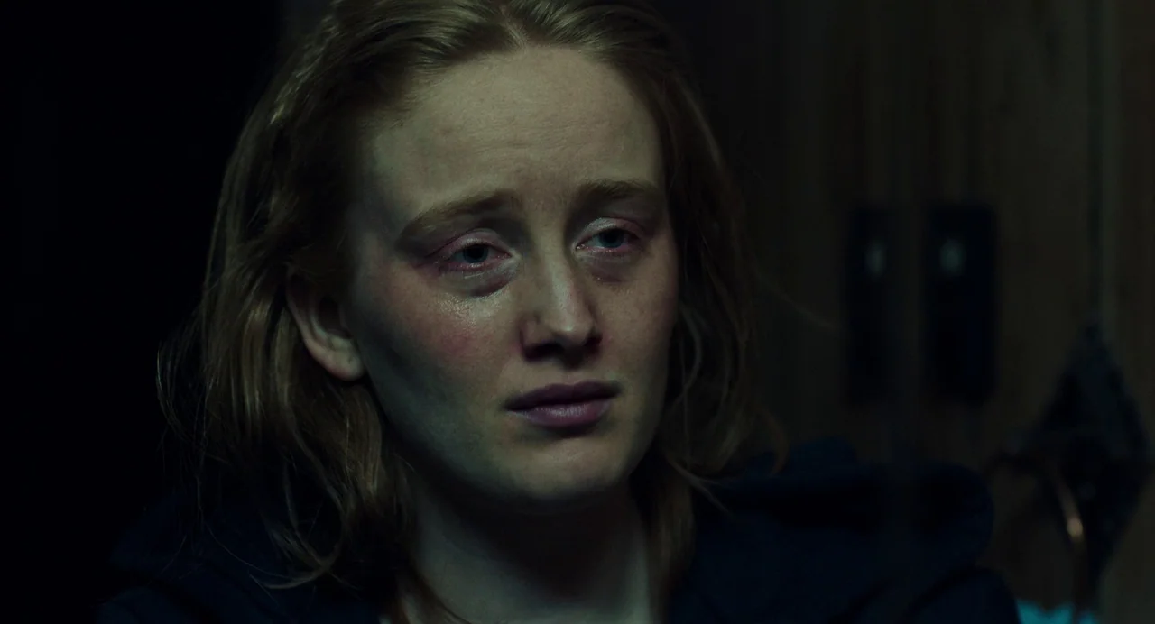 My Animal (2023) movie still: closeup — A dimly lit indoor space with a dark, indistinct background. A young woman looks off-camera with visible…