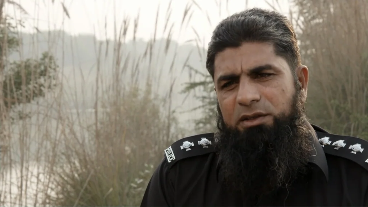 A Girl in the River: The Price of Forgiveness (2015) movie still: medium — A man in a black police uniform stands in an outdoor area with tall…