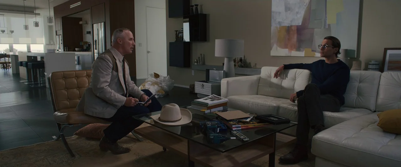 Intrusion (2021) movie still: wide — A minimalist modern living room features a white leather couch and glass coffee table. Two men sit in…