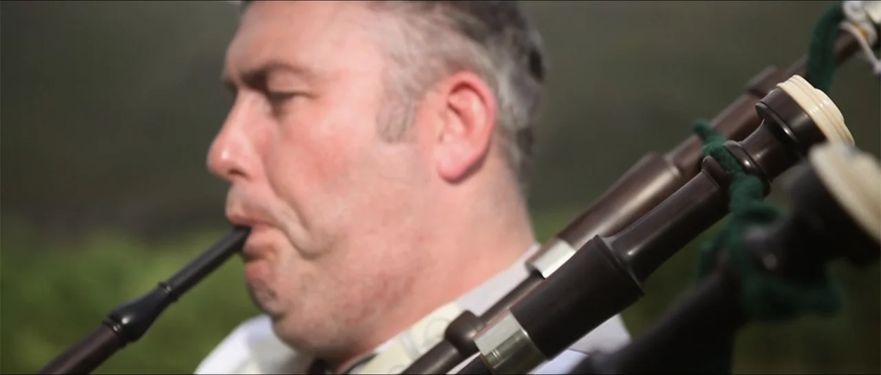 Garnet’s Gold (2014) movie still: closeup — A man in a light shirt plays the bagpipes outdoors against a blurred green backdrop. The focus remains…