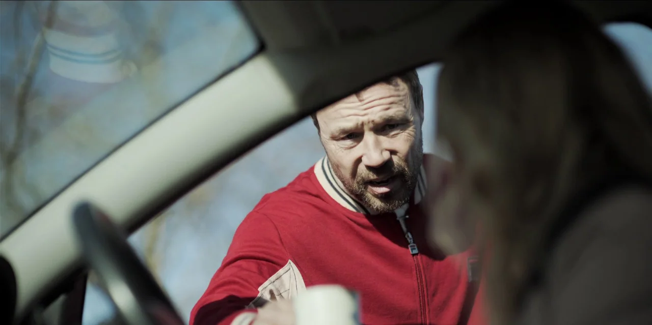 Help (2021) movie still: medium — A man stands outside a car, leaning through the open window to talk. The interior is partially obscured by the…