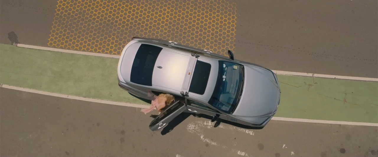 Anyone But You (2023) movie still: wide — An asphalt road features a green bike lane and a yellow hexagonal pattern. A woman exits a silver sedan…