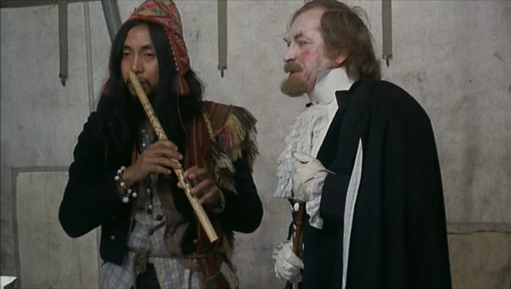 The Enigma Of Kaspar Hauser (1974) movie still: medium — Two men stand inside a canvas tent with plain off-white fabric walls. One man plays a…