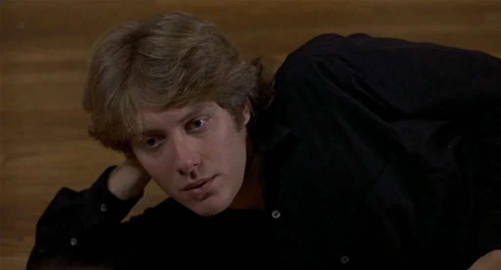 Sex, Lies And Videotape movie still: closeup — A man lies on a wooden floor in a dimly lit space. He rests his head on his hand while looking toward…