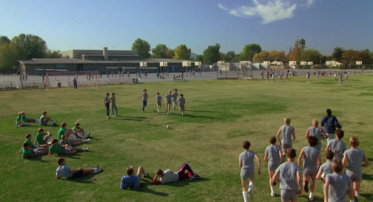 The Karate Kid (1984) movie still: wide — A sprawling grassy athletic field sits in front of a low-rise school building. Dozens of students in…