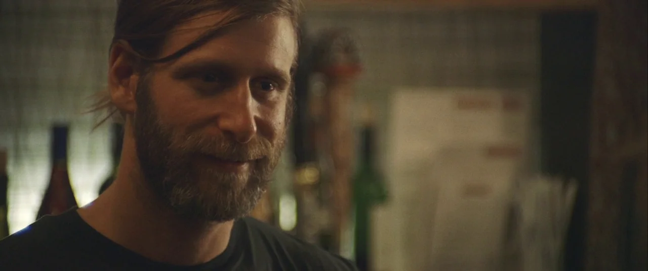 Always Shine (2016) movie still: closeup — A dimly lit bar setting with blurred bottles visible in the background. A man with a beard looks toward…