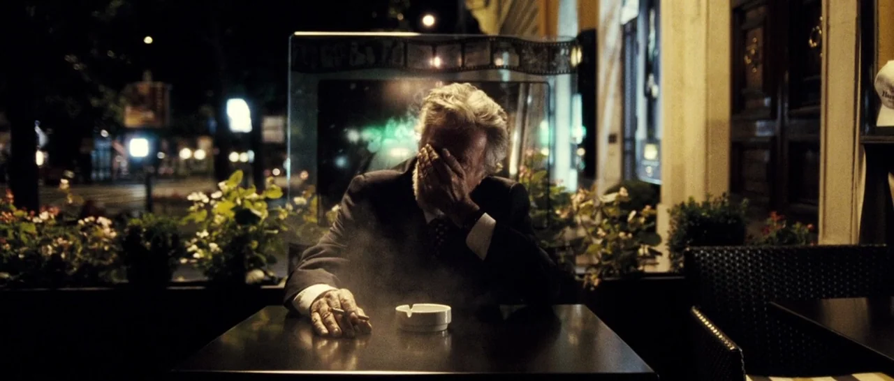 The Great Beauty (2013) movie still: medium — A night view of an outdoor cafe terrace at a wooden table. An older man in a dark suit rests one hand…