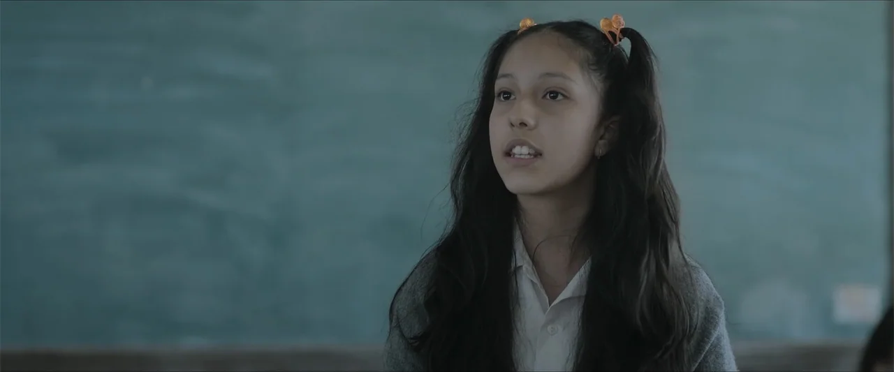 Radical (2023) movie still: medium — A student stands in front of a green chalkboard in a quiet school room. The young girl speaks while facing…