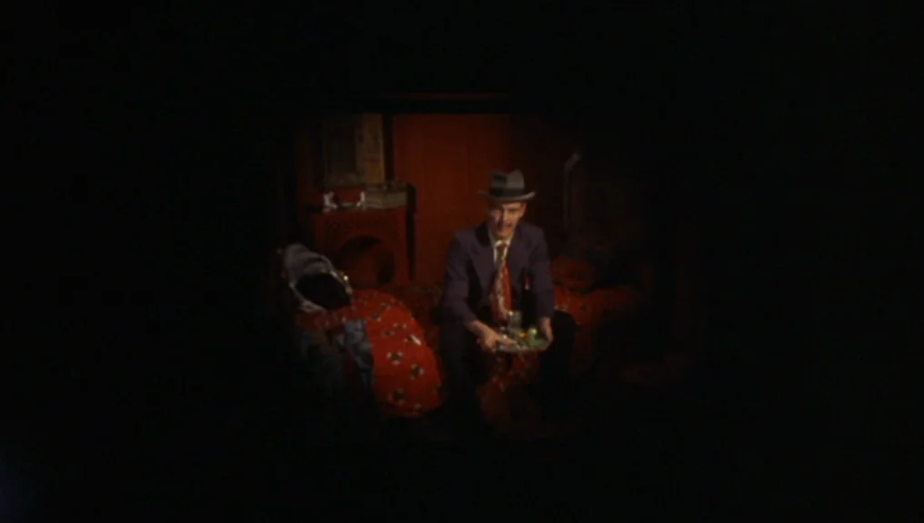 Performance (1970) movie still: medium — A man sits on a bed inside a dark room with red patterned bedding. He holds a tray while facing the camera…