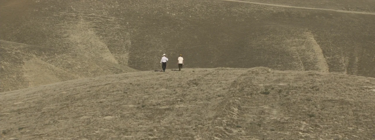 Entertainment (2015) movie still: wide — Two men walk across a barren, dry, rolling hill landscape under a pale sky. The vast, dusty terrain…