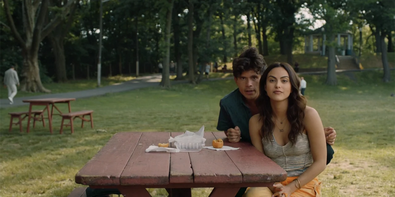 Música (2024) movie still: medium — A green park setting features a red picnic table in the foreground. A young man and woman sit behind the table…