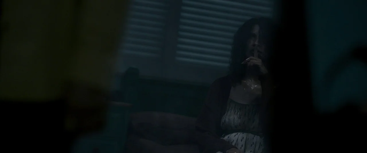 Talk To Me (2022) movie still: medium — A dimly lit room at night features closed window blinds in the background. A woman sits on a surface while…