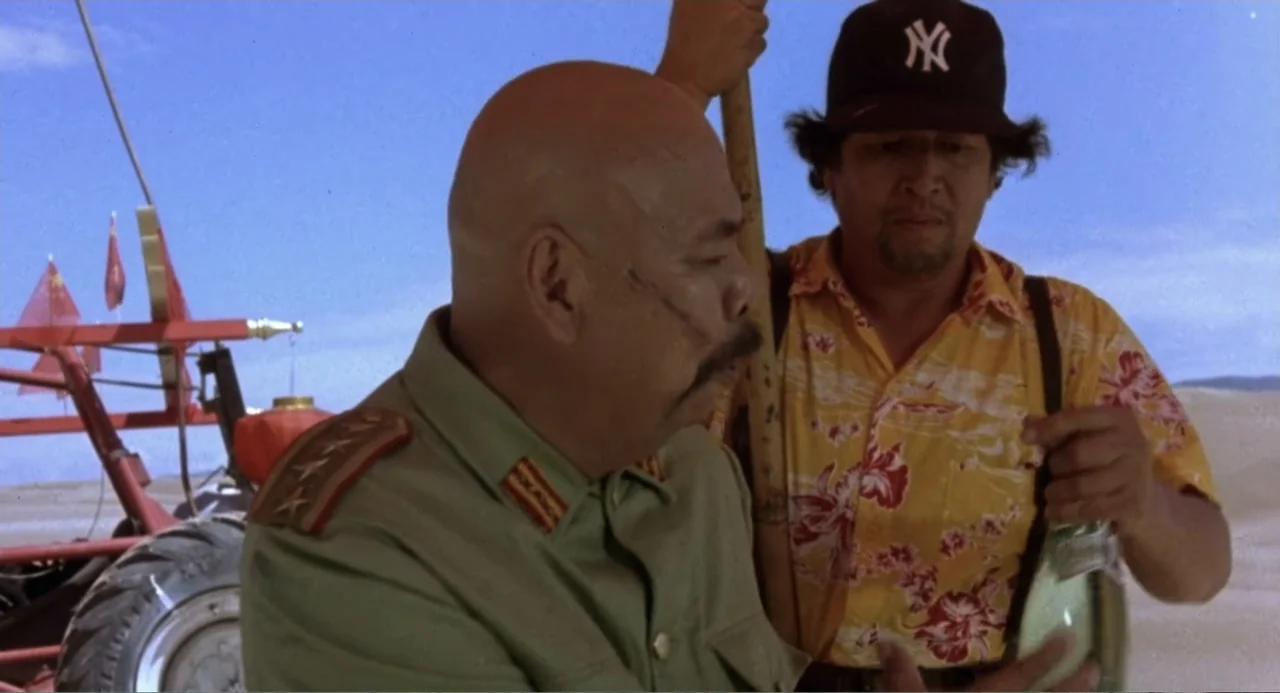 Firewalker (1986) movie still: medium — A flat sandy desert landscape under a clear bright blue sky. A bald officer in military uniform stands…