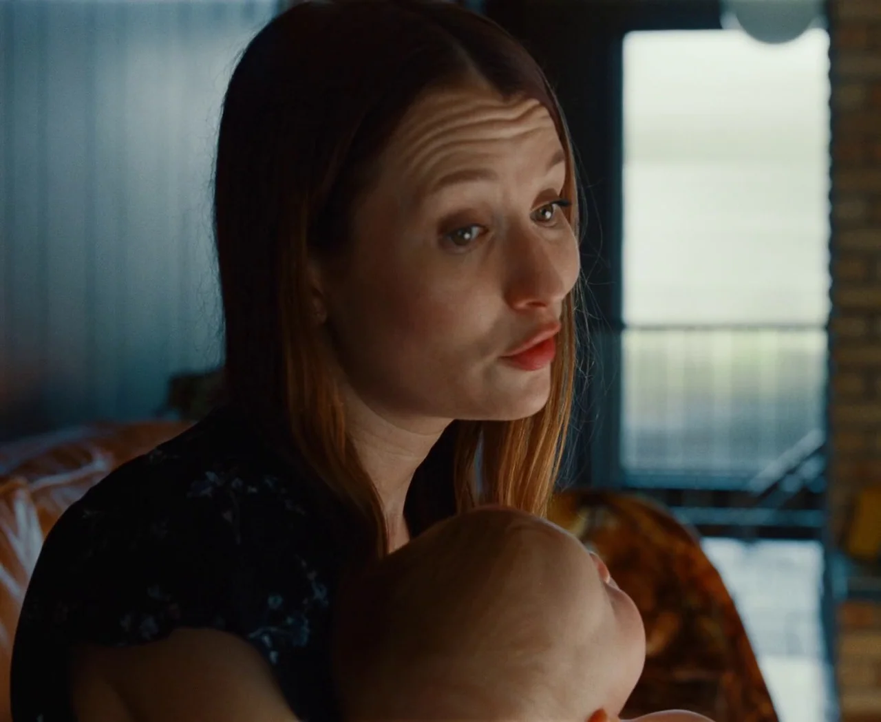 Monica (2022) movie still: closeup — A woman holds an infant inside a room with a blurred window backdrop. The woman looks away from the camera as…