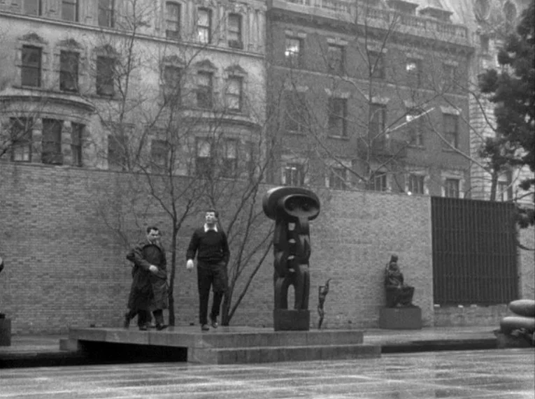 Shadows (1958) movie still: wide — A stone platform in a public courtyard features a large abstract totem-like statue. Two men walk across the wet…