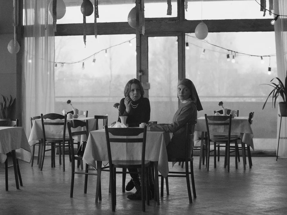 Ida (2013) movie still: medium — A spacious room with large windows features multiple tables draped in white cloths. Two women sit together at a…