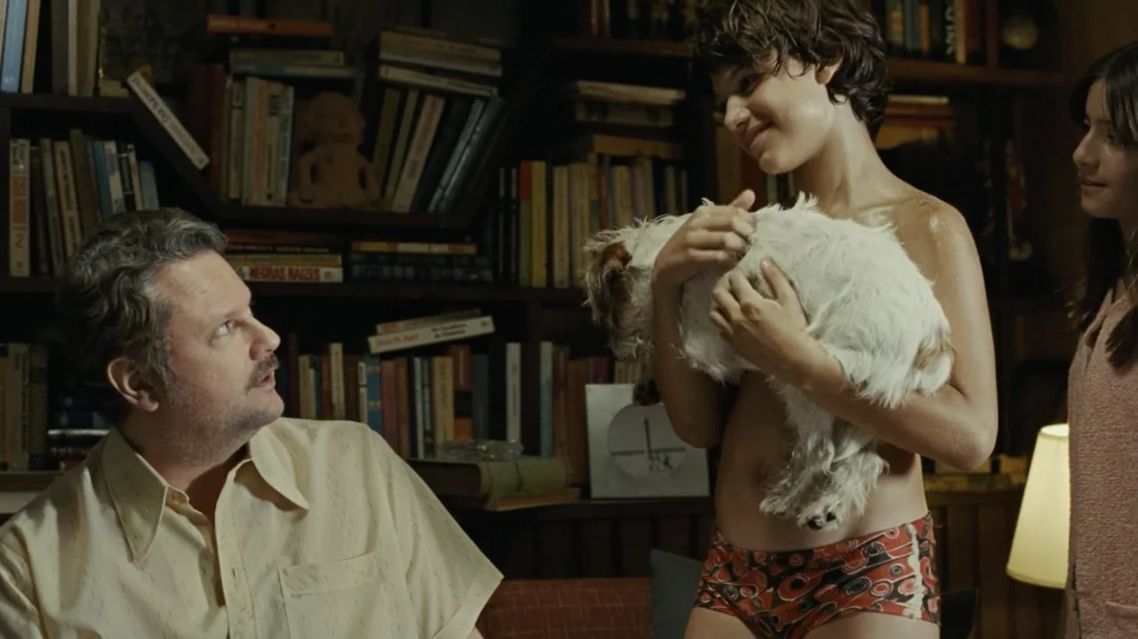 I'm Still Here (2024) movie still: medium — A book-lined study features shelves packed with various volumes and a small sculpture. A man sits while…