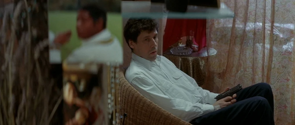 The Crying Game (1992) movie still: medium — A man sits in a wicker chair within a patterned, cluttered domestic interior. He holds a dark…