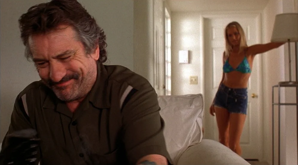 Jackie Brown movie still: medium — A living room interior features a man sitting near a white sofa. A woman in a bikini and denim shorts stands in…