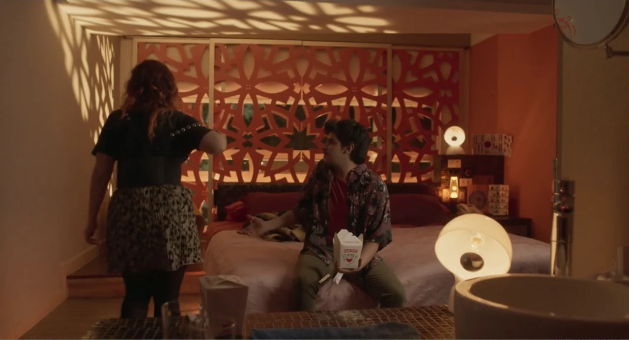Casando a mi Ex (2023) movie still: medium — A hotel room features a bed positioned in front of a geometric orange room divider. A man sits on the…