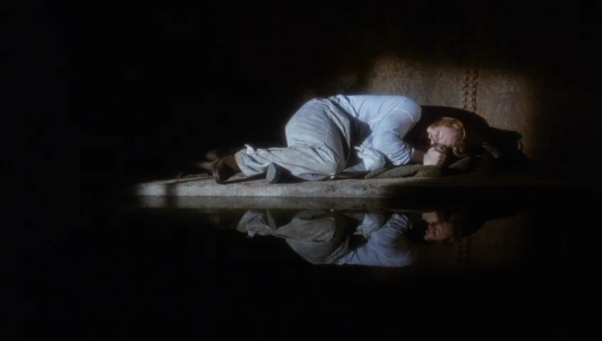 Edward II (1991) movie still: wide — A dark subterranean concrete structure with water reflecting the figure on a ledge. A man lies curled up in the…