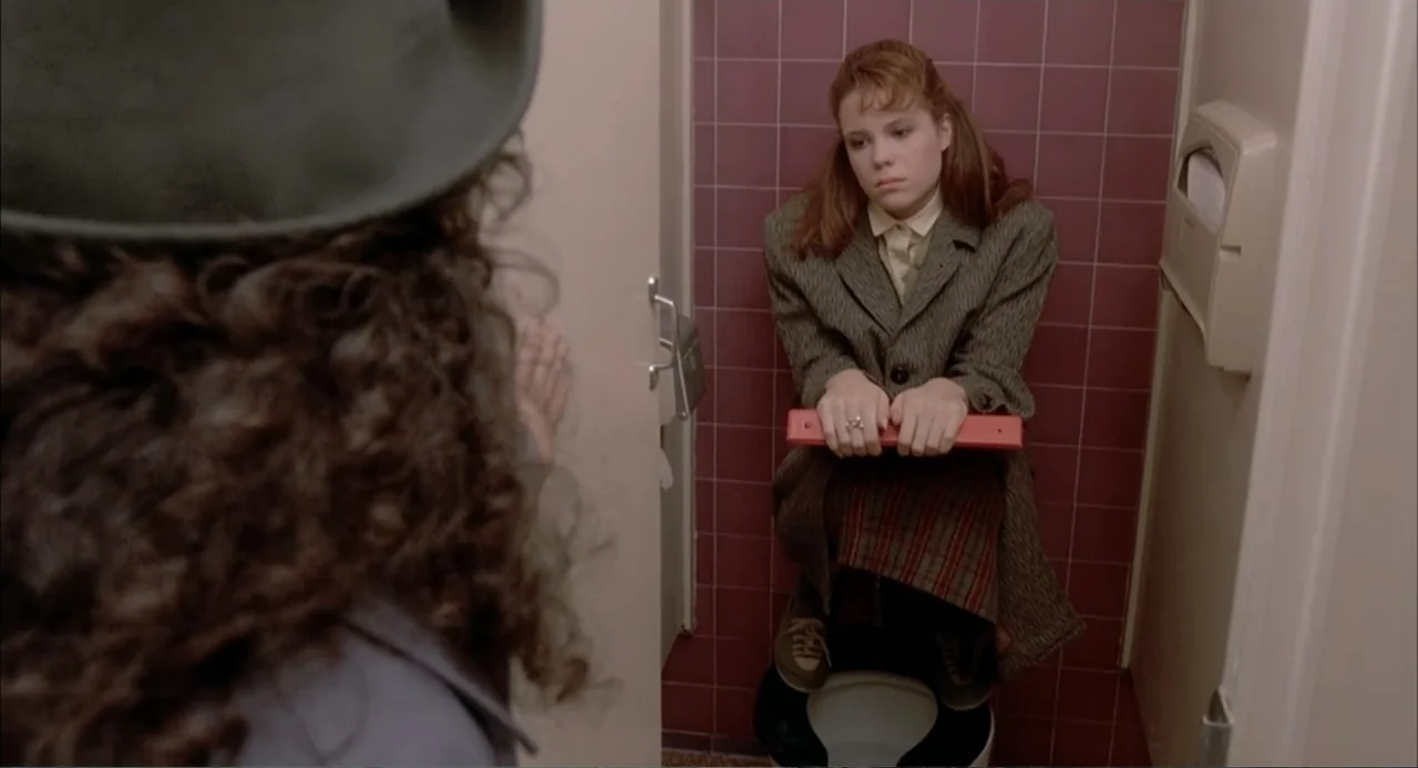 Teen Witch (1989) movie still: medium — A cramped bathroom stall features dark maroon tiled walls and a beige partition door. A teen girl in a tweed…
