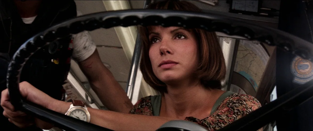 Speed (1994) movie still: closeup — The interior of a bus showing the driver seated behind the large steering wheel. The woman holds the wheel while…