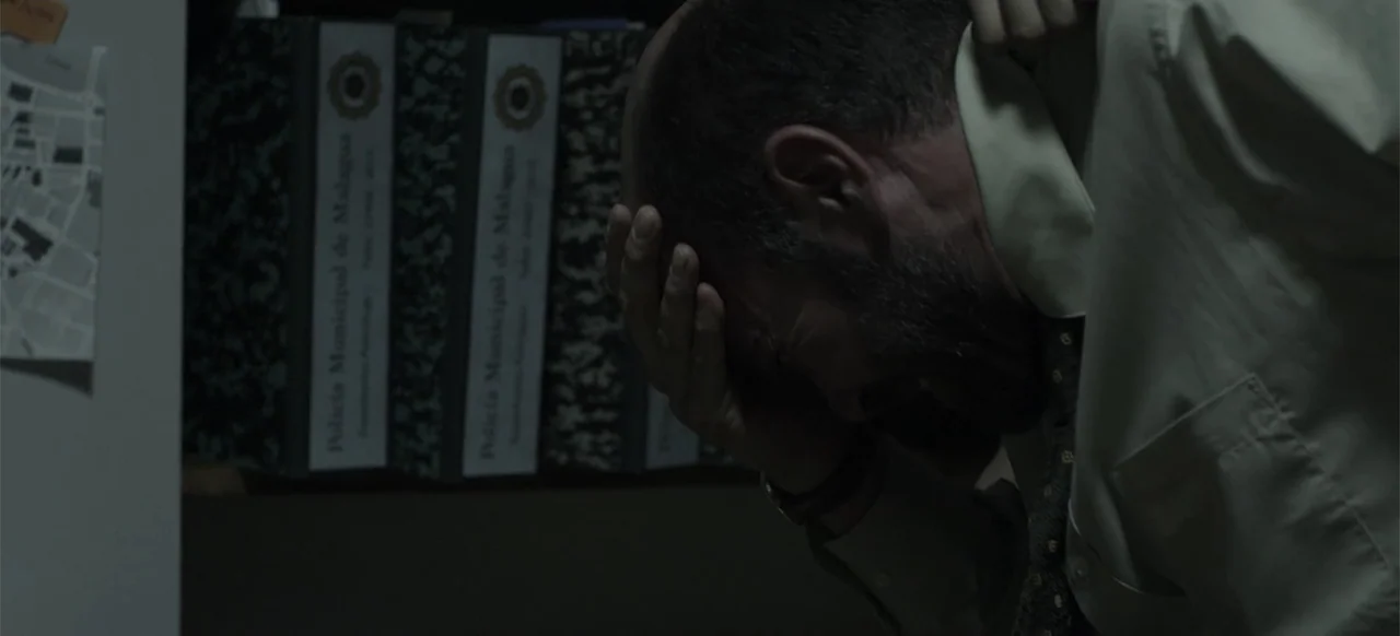 About the Deaths (2016) movie still: closeup — A dimly lit office features several municipal binders on a shelf in the background. A man in a dress…