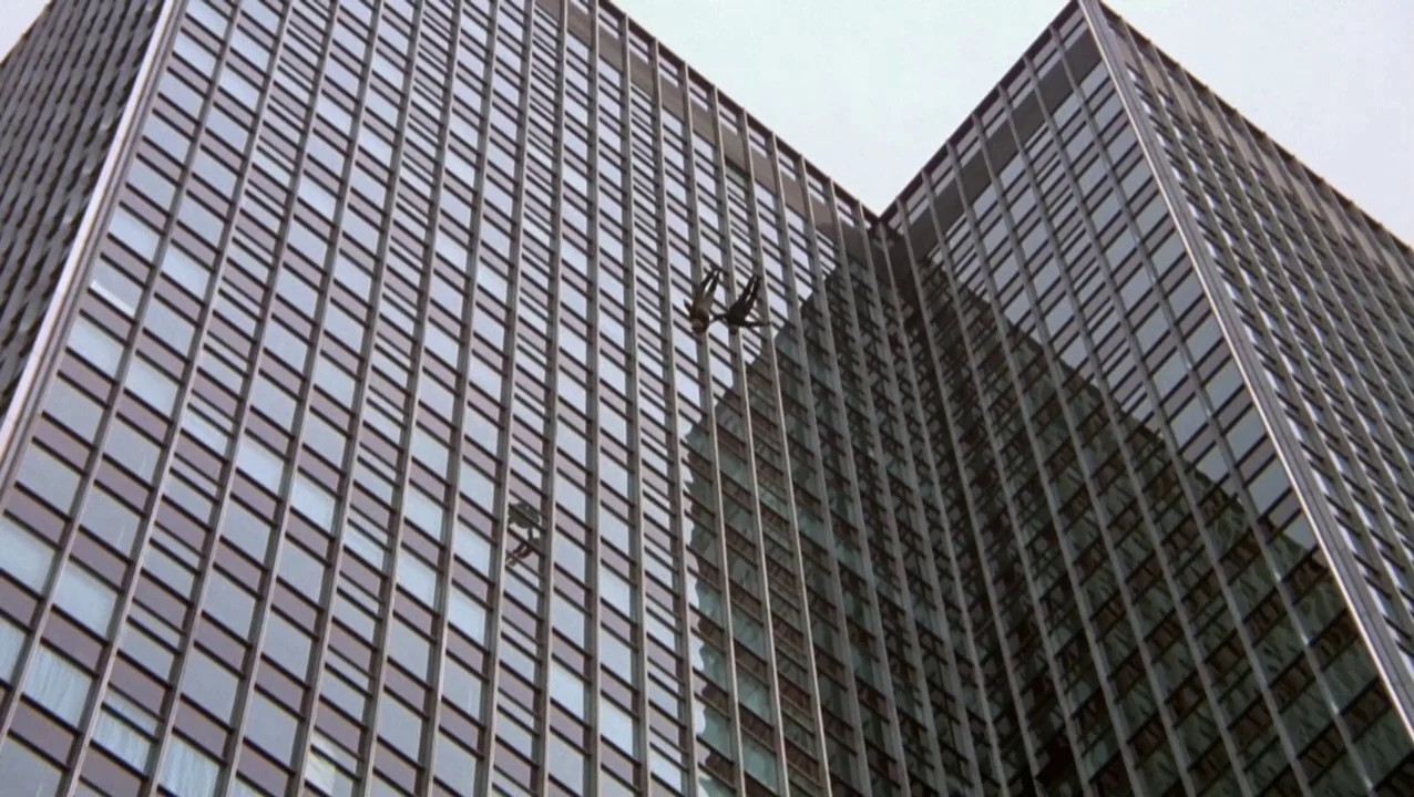 O Lucky Man! (1973) movie still: establishing — A massive, angular glass skyscraper looms against a bright, overcast sky. Two small human figures…