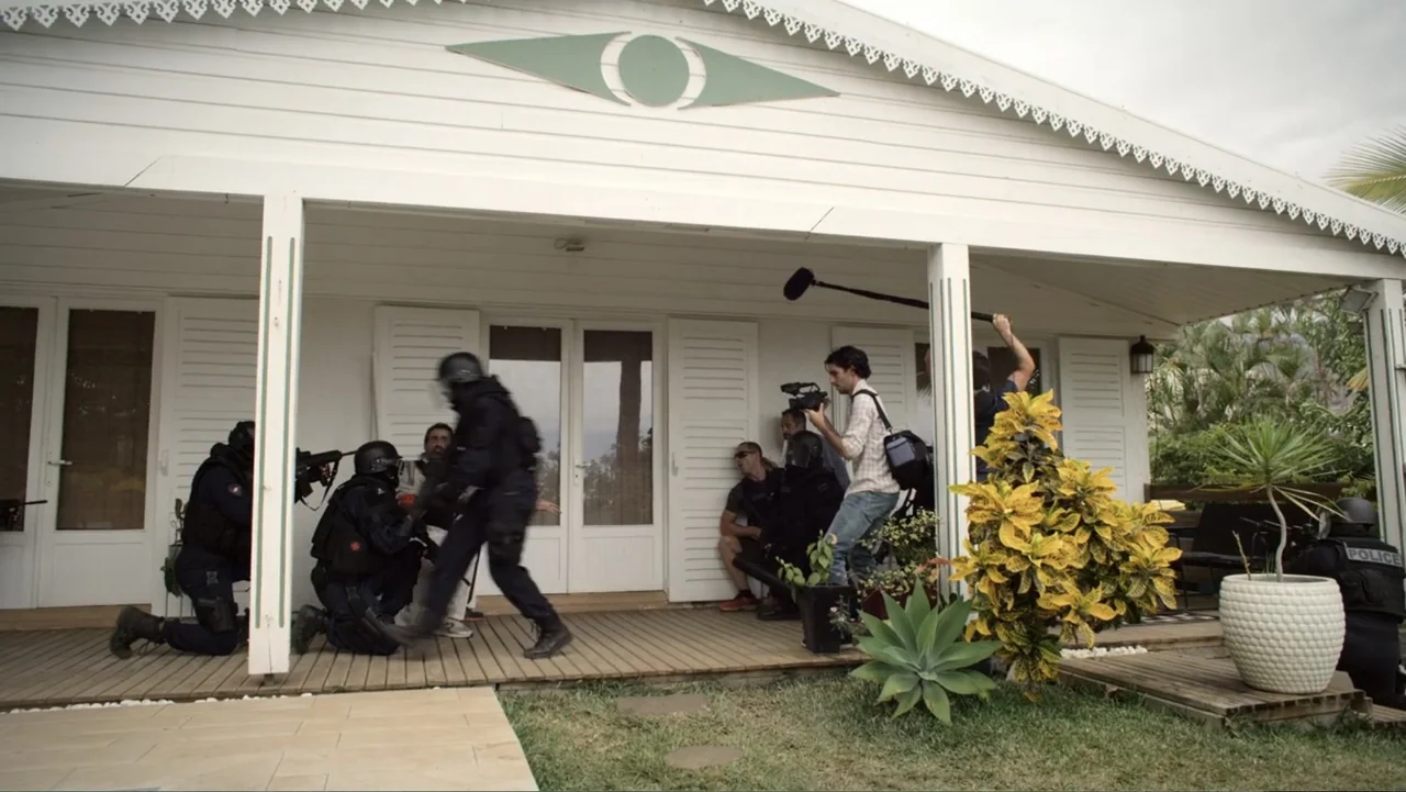 Unknown #1 movie still: wide — A white house exterior features a wooden porch occupied by tactical police and film crew. One camera operator stands…