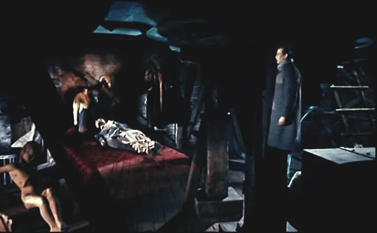 Mill of the Stone Women (1960) movie still: wide — A dark, cluttered attic space features heavy wooden rafters and uneven lighting. A man stands at…