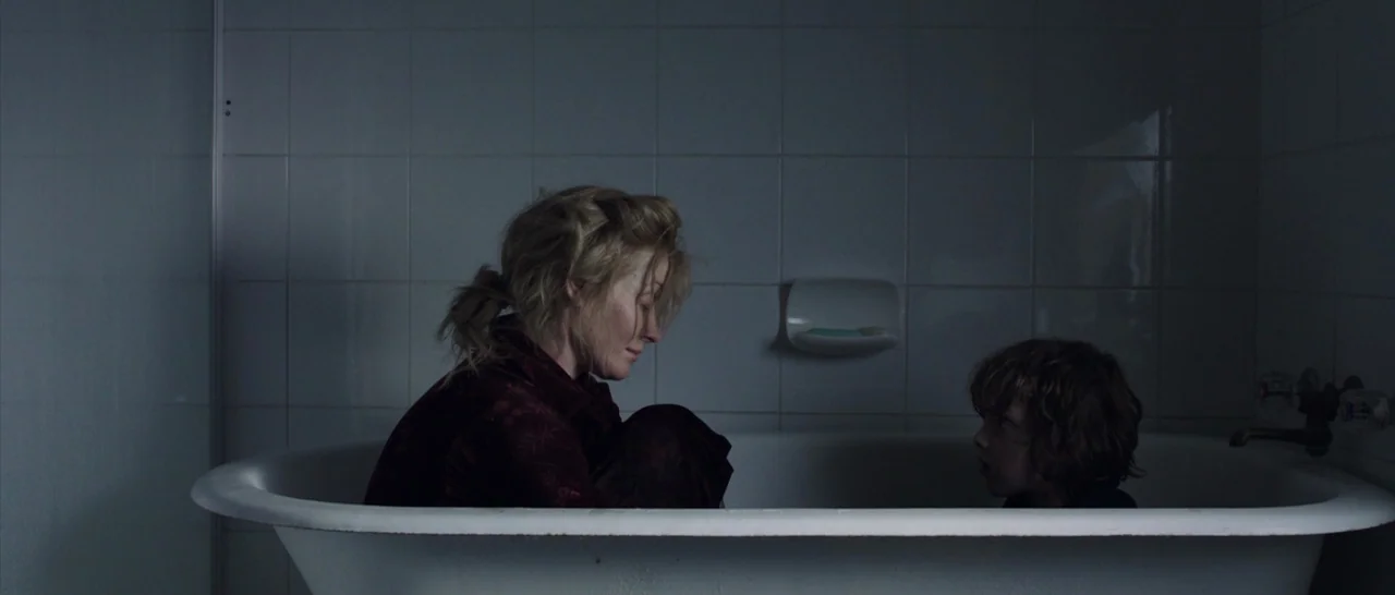 The Babadook (2014) movie still: medium — A dimly lit room features white tiled walls and a porcelain bathtub. A woman and young boy sit in the tub…