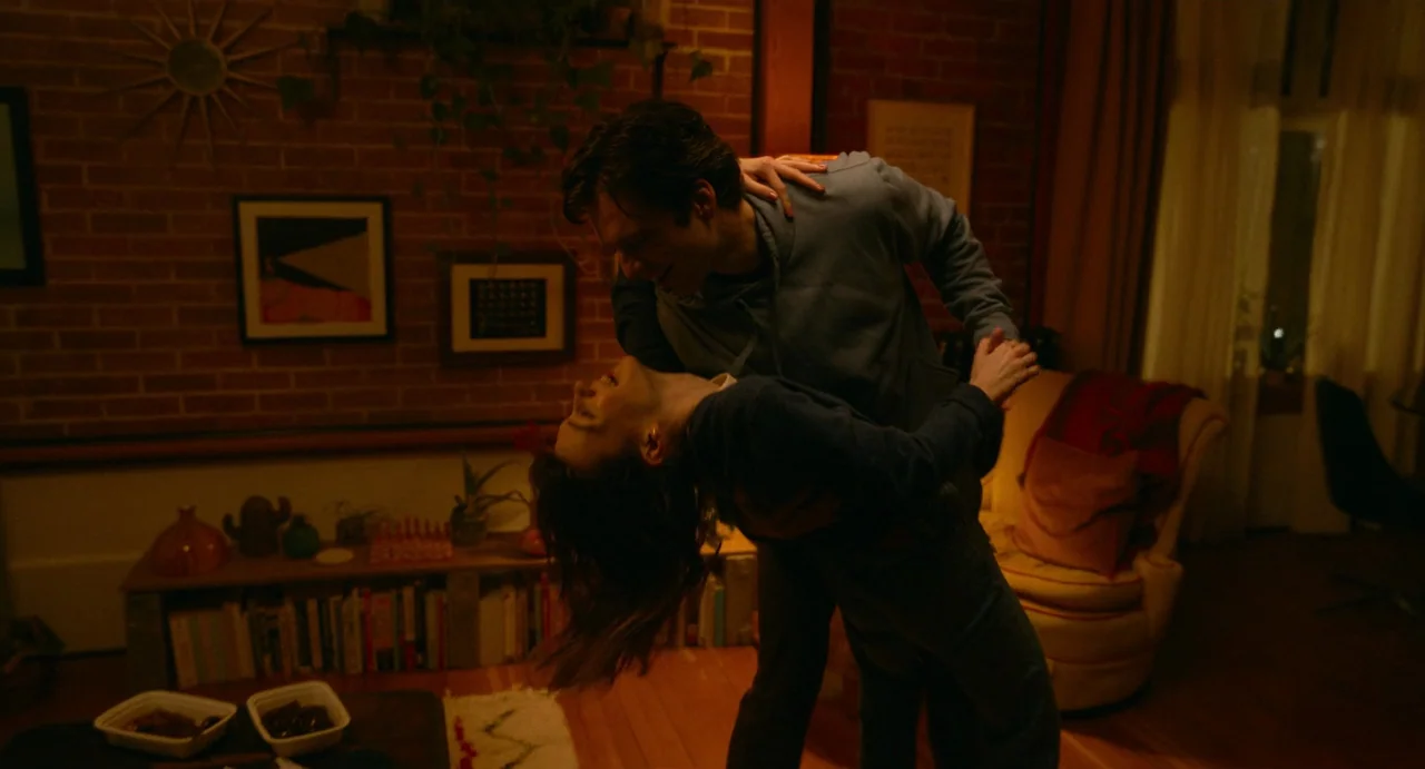 Fresh (2022) movie still: medium — A cozy room with exposed brick walls and a bookshelf beneath a window ledge. A couple stands in the center of the…
