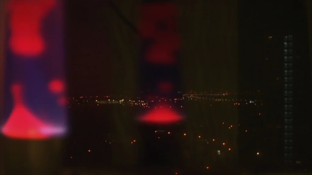 Red Road (2006) movie still: wide — A view through an interior window at night showing a distant urban sprawl. Two red lava lamps occupy the…