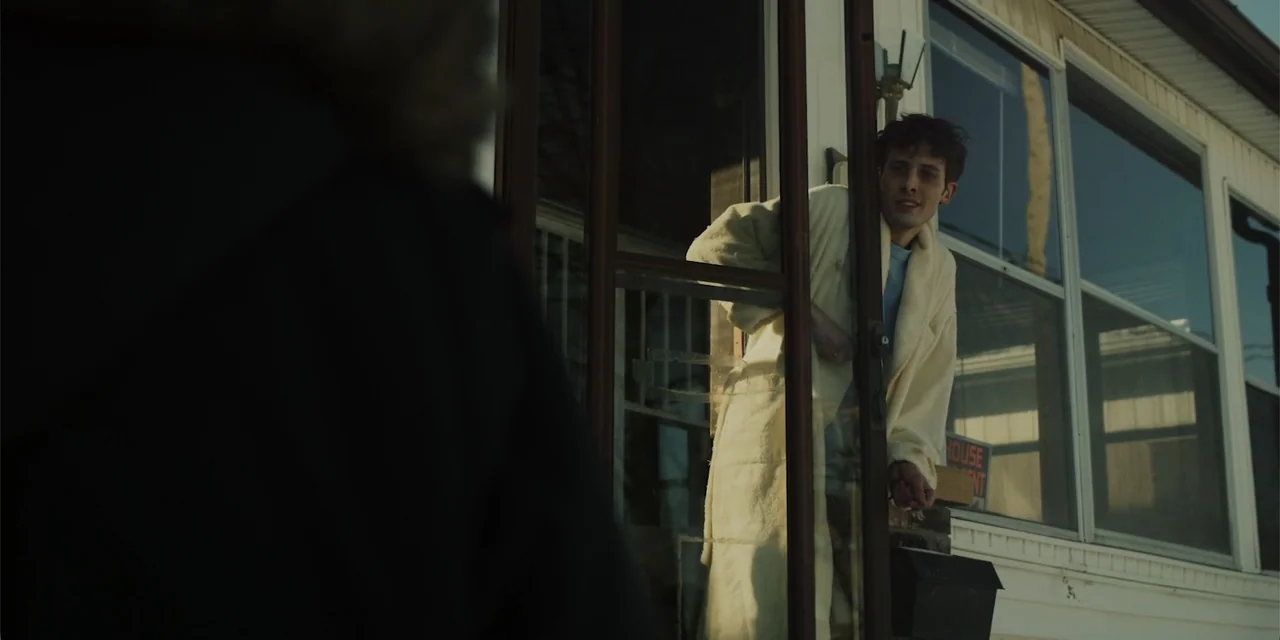 Lobos (2019) movie still: medium — The exterior of a residential home shows a screen door opening onto a porch. A young man wearing a bathrobe…