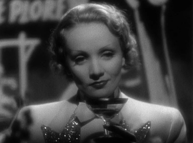 Blonde Venus (1932) movie still: closeup — A dark, dimly lit environment features a woman positioned in the foreground. The subject holds a cocktail…