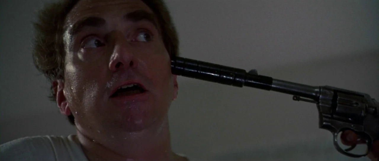 Thunderbolt & Lightfoot (1974) movie still: closeup — A man looks up at a firearm pressed firmly against his cheekbone. The background remains dark…