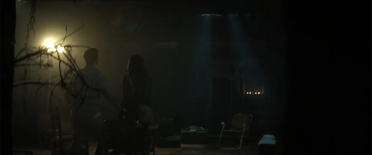 Truth or Dare (2018) movie still: wide — A dark interior space features scattered furniture including folding chairs and a plastic cooler. A man and…