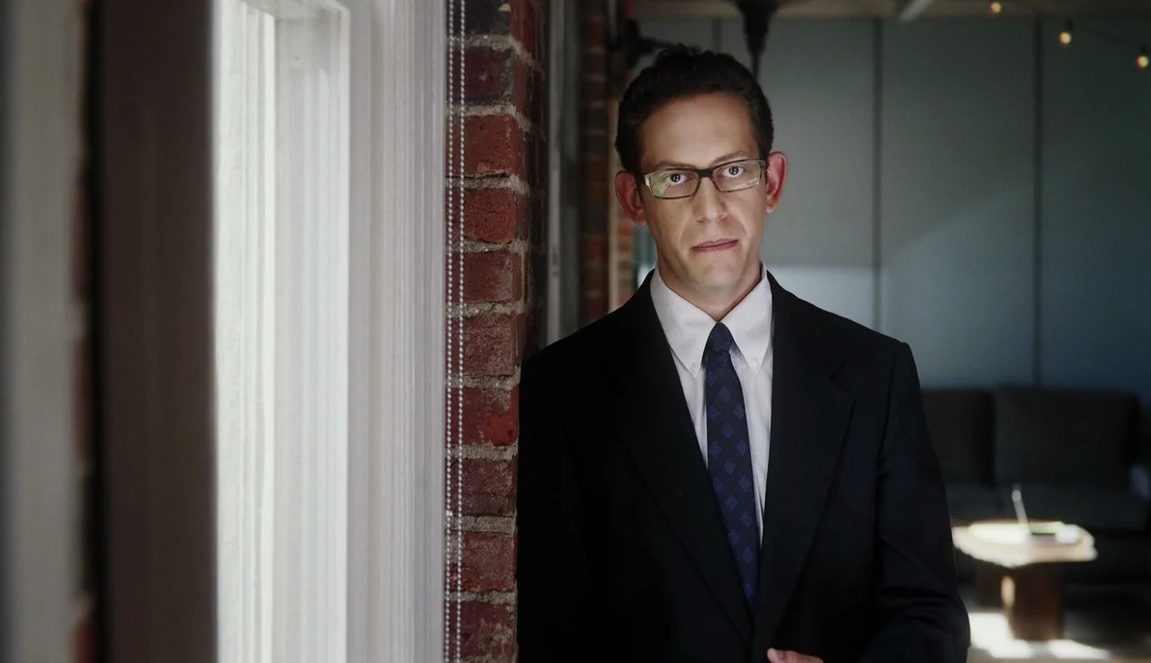13th (2016) movie still: medium — A man stands indoors next to a brick pillar and a large window. He is positioned in a room with a sofa and coffee…