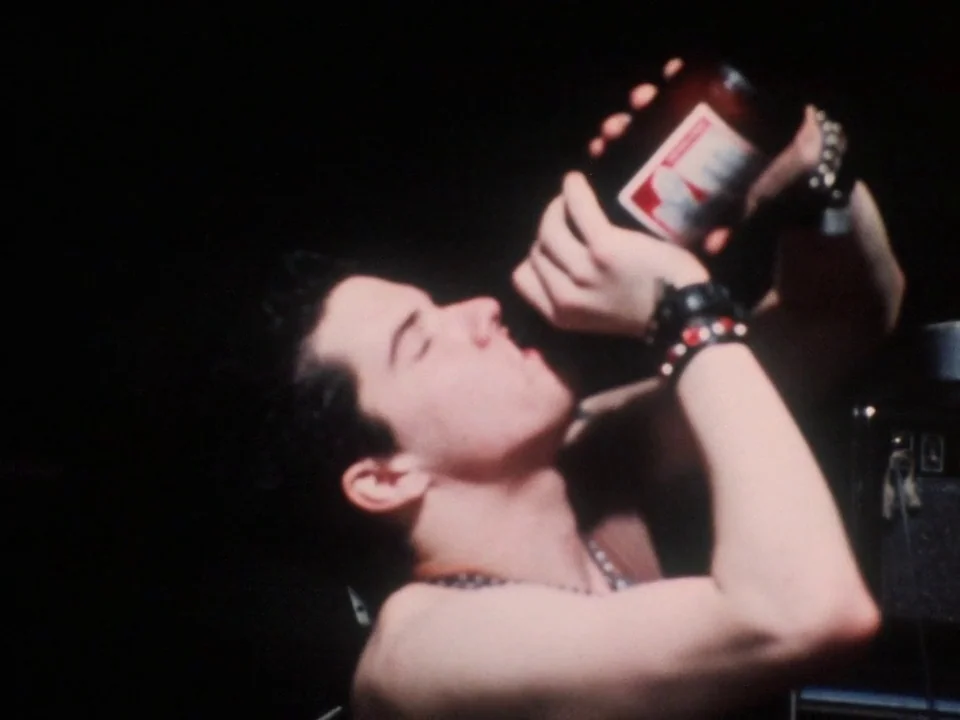 Decline of Western Civilization (1981) movie still: closeup — A dark, minimally lit environment frames a young man in profile. He tilts his head…