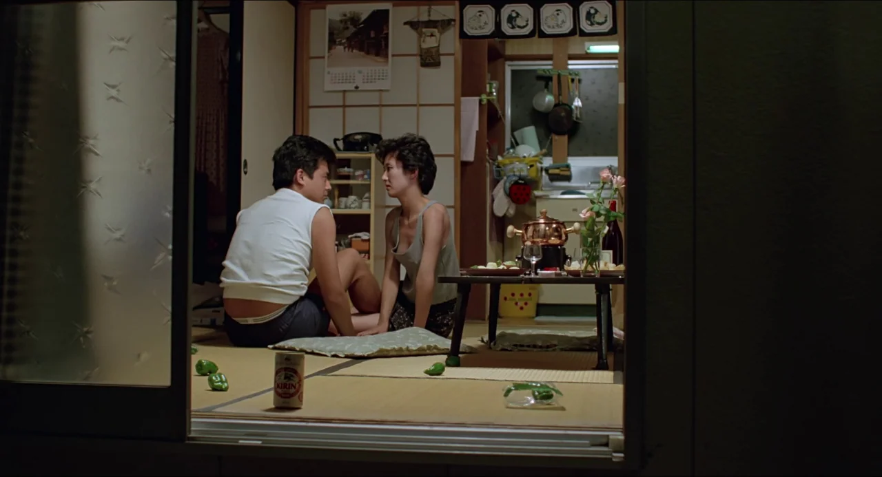 Typhoon Club (1985) movie still: wide — A Japanese apartment room features straw mat flooring and a low wooden dining table. A man and a woman sit…