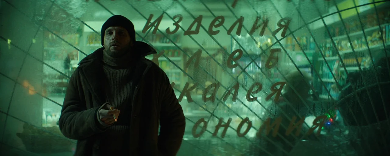 Petrov’s Flu (2021) movie still: medium — A man stands outside a store front behind a glass window with Cyrillic text. Reflections and patterns are…