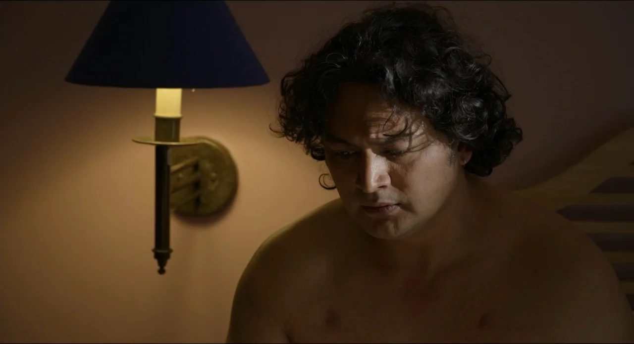 The Beauty and the Moody (2022) movie still: medium — A dimly lit room with a wall-mounted lamp providing a soft light source. A man sits shirtless…
