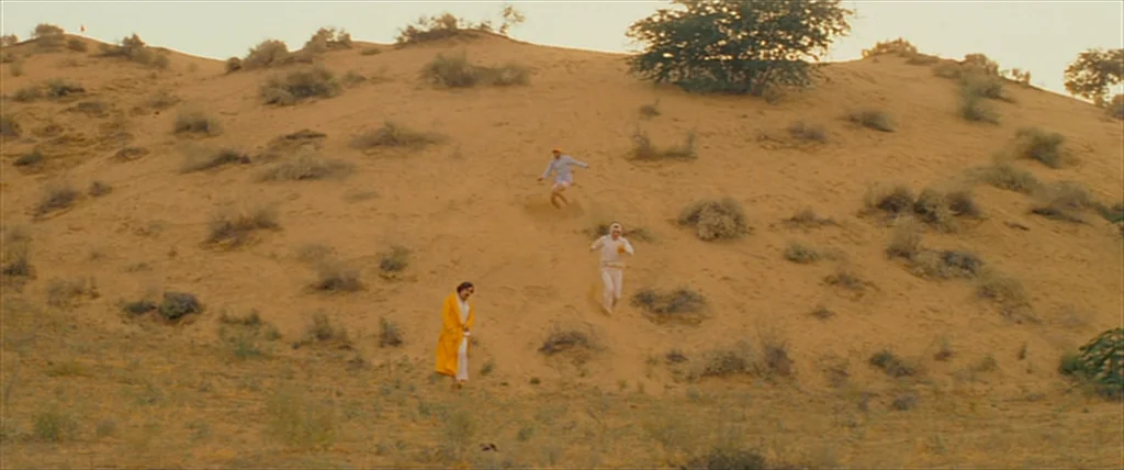 The Darjeeling Limited (2007) movie still: wide — A vast, arid desert landscape features rolling sand dunes dotted with sparse, dry shrubs. Three…