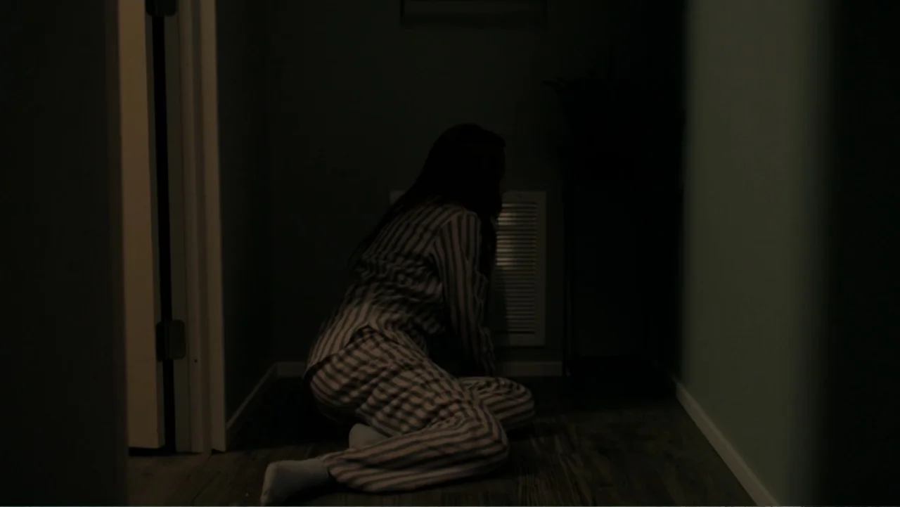 Sightless (2017) movie still: medium — A dimly lit indoor hallway with a wooden floor and dark teal walls. A person in striped pajamas sits on the…