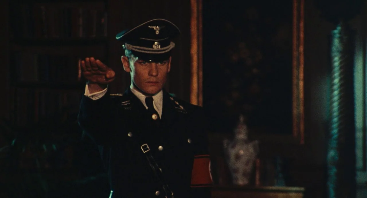 The Damned (1969) movie still: medium — A dark, wood-paneled room contains shelves of books and an ornate framed portrait. A young man wearing a…